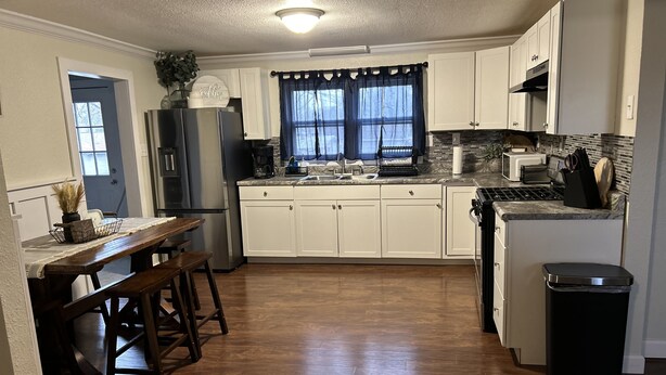 Una cocina con armarios blancos, una mesa de comedor de madera, una estufa integrada y un cubo de basura.
