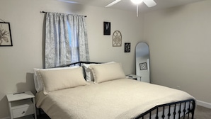 Una cama bien hecha con edredón blanco y almohadas, una mesita de noche con lámpara, un espejo fijado a la pared y un ventilador de techo.