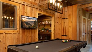 Une table de billard avec une seule boule blanche, des murs en bois, un tableau représentant un paysage encadré et une étagère contenant des ustensiles de cuisine.