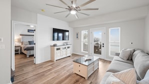 Ein geräumiges Wohnzimmer mit Holzboden, Deckenventilator, Flachbildfernseher, Glastisch und grauem Sofa.