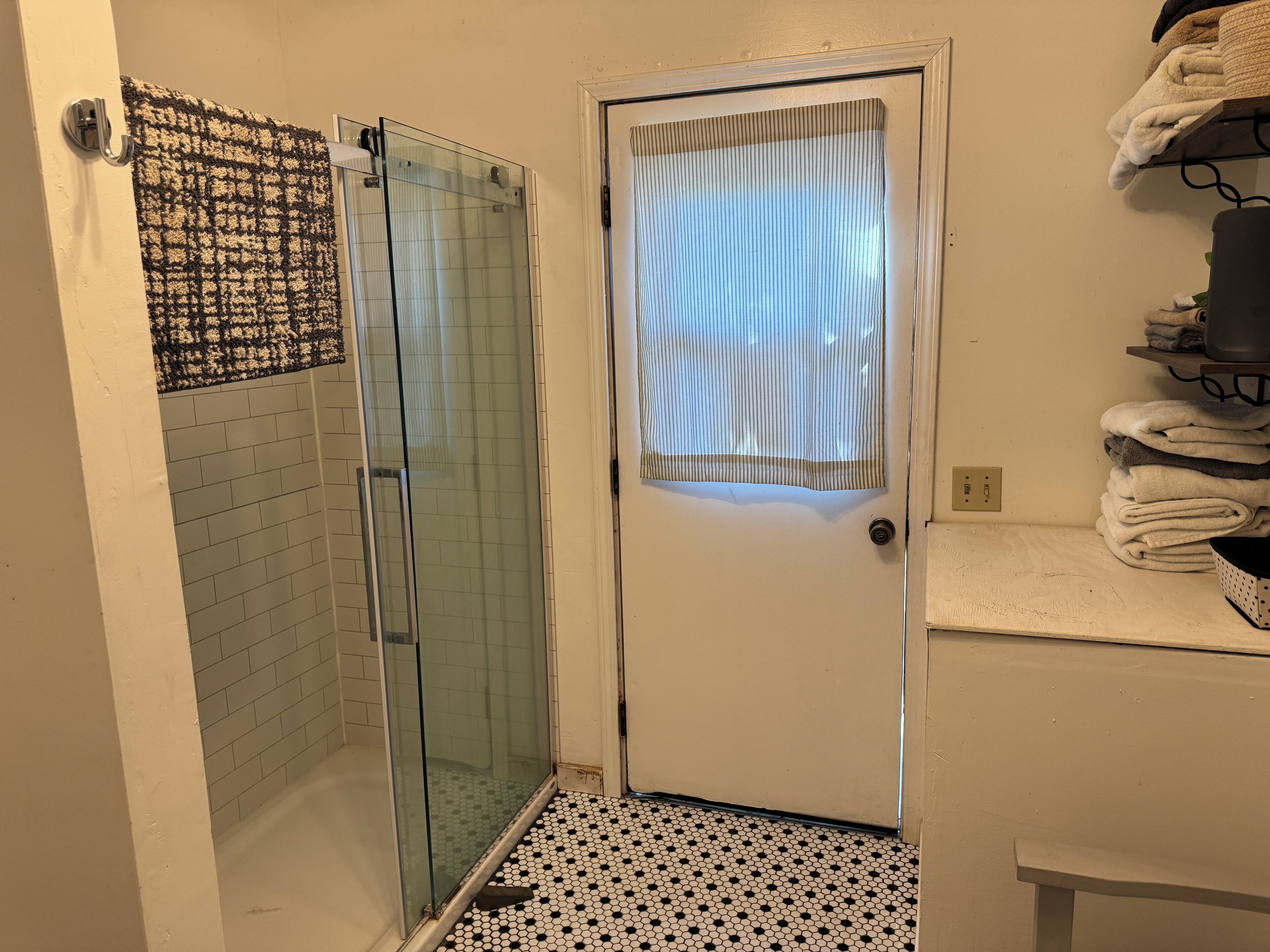 Um banheiro com box de vidro, piso ladrilhado, porta branca com janela e porta-toalhas com toalhas dobradas.