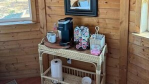 A wooden cabin interior with a small table holding a coffee maker, cups, and a basket of snacks. A mirror hangs on the wall above the table. - Stay on the farm - 1 bedroom cottage with deck outside of Davis (Davis)