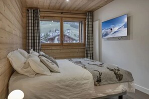 Quarto com teto de madeira, uma janela grande com cortinas, cama com manta estampada e televisão exibindo uma paisagem nevada.
