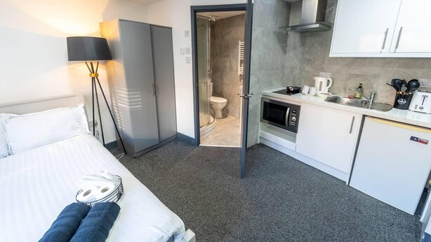 A hotel room with a bed, a bedside lamp, a wardrobe, a bathroom with a toilet and shower, and a kitchenette with a sink and stove. - Captivating 1-bed Studio in Birmingham (Birmingham)
