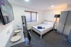 A hotel room with a bed, a desk with a chair, a mirror, a radiator, a window with blinds, a floor lamp, and a wardrobe. - Captivating 1-bed Studio in Birmingham (Birmingham)