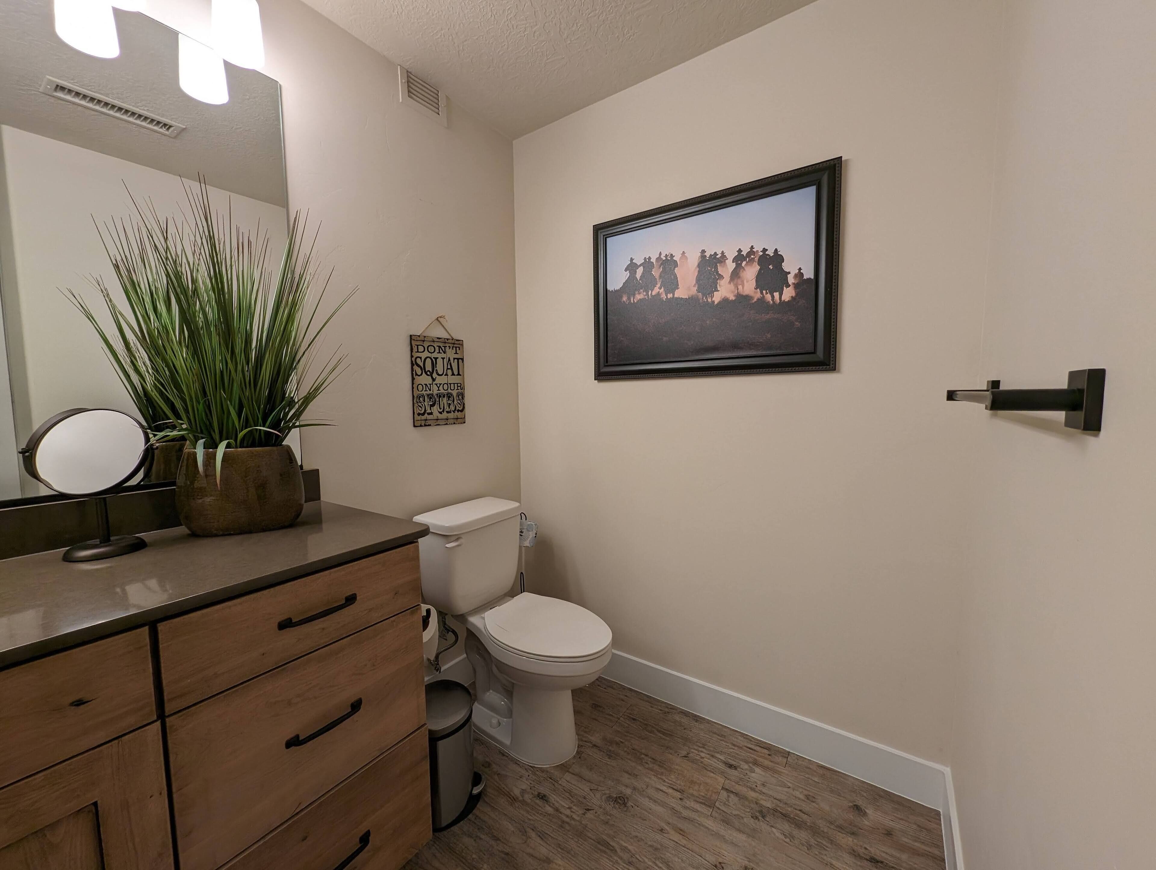 Une salle de bain avec un meuble-lavabo en bois, un miroir, une plante en pot, des toilettes et un tableau encadré au mur.