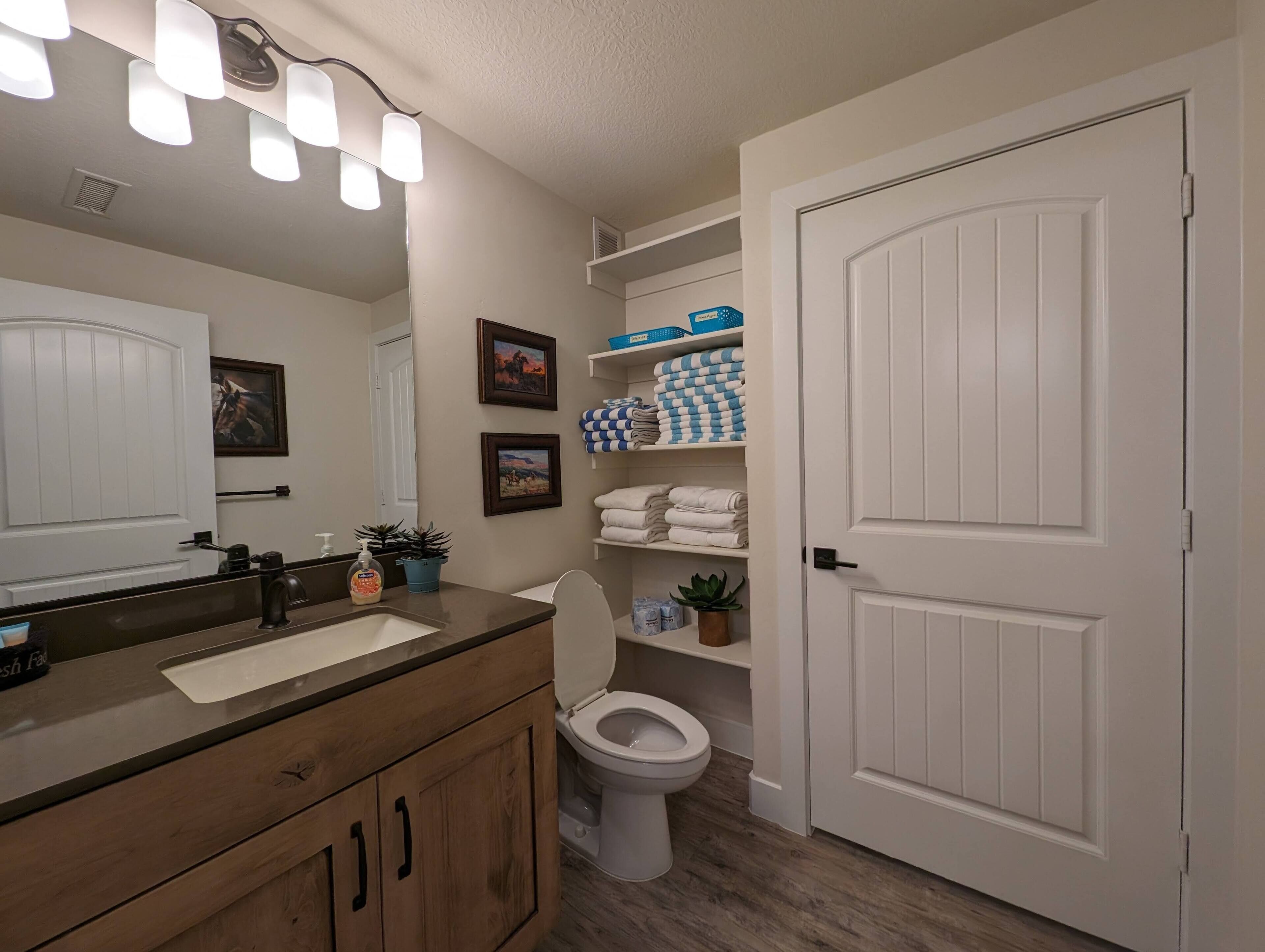 A bathroom with a wooden vanity, a white toilet, and shelves with folded towels.