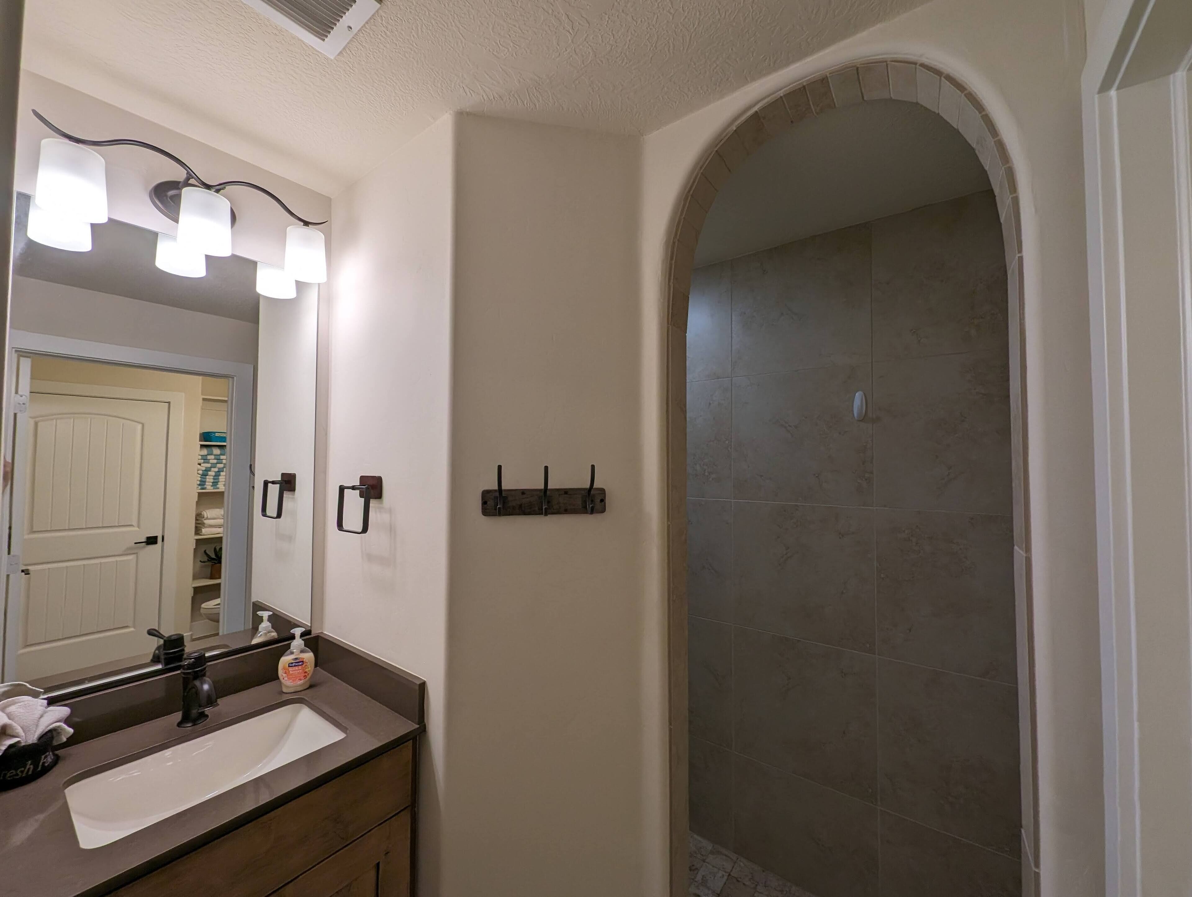 Une salle de bain équipée d’un lavabo, d’un miroir et de luminaires. On y trouve également un porte-serviettes et un espace douche dont les murs sont recouverts de carreaux.