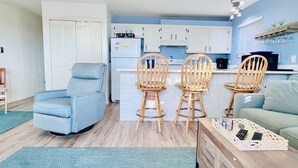 A living room with a blue armchair, two wooden chairs, a wooden coffee table with remote controls, and a kitchen in the background. - Stunning 2BR/2BA Bayfront Retreat (Ocean City)