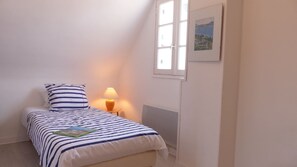 Ein kleines, ordentlich eingerichtetes Schlafzimmer mit einem Einzelbett, einem Nachttisch mit Lampe, einem Heizkörper und einem Fenster mit Blick ins Grüne.
