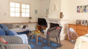 Ein gemütliches Wohnzimmer mit einer Couch, einem Korbsessel, einem kleinen Tisch mit Obst, einem Fernseher und einem Kamin.
