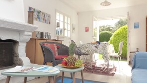 A cozy living room with a fireplace, wicker furniture, a dining area with a table and chairs, and a view of a garden through an open door.