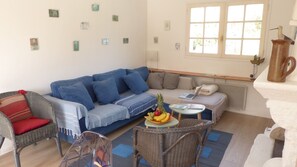 A cozy living room with a blue sofa, a wicker chair, a small table with fruits, and a window with a view.