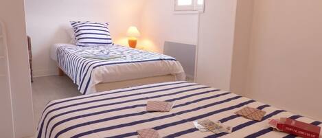A room with two beds, one with a striped bedspread and the other with a plain one. There is a lamp on the nightstand beside the bed with the striped bedspread.