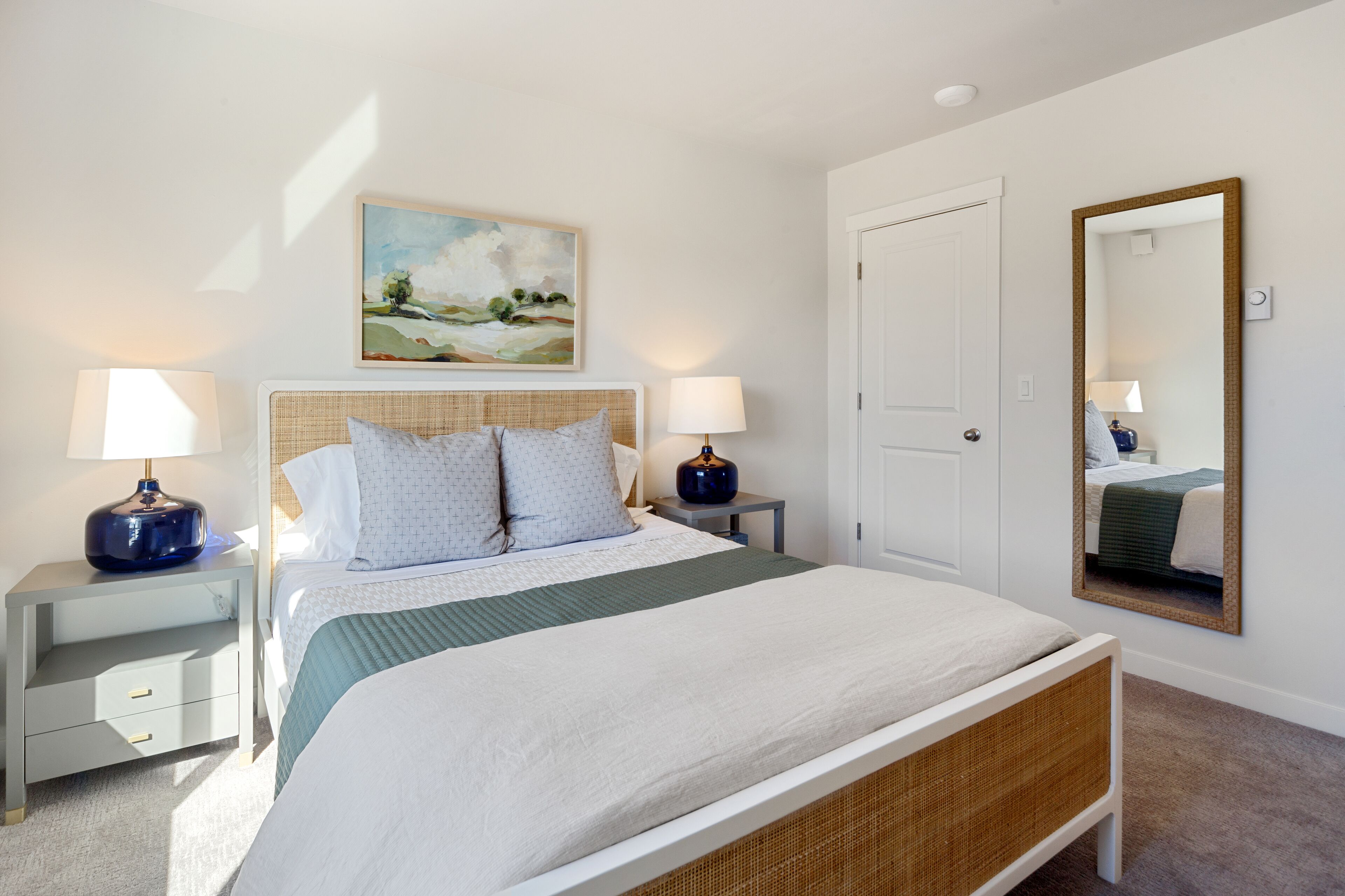 Un dormitorio ordenado con una cama grande, dos mesitas de noche con lámparas, un cuadro en la pared, una puerta y un espejo.