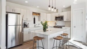 A modern kitchen with white cabinetry, stainless steel appliances, a central island with stools, and pendant lighting.