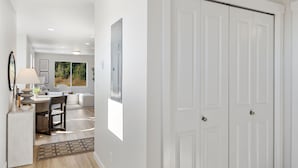 A hallway with a white door, a desk with a lamp, and a view of a living area with a couch and a window.