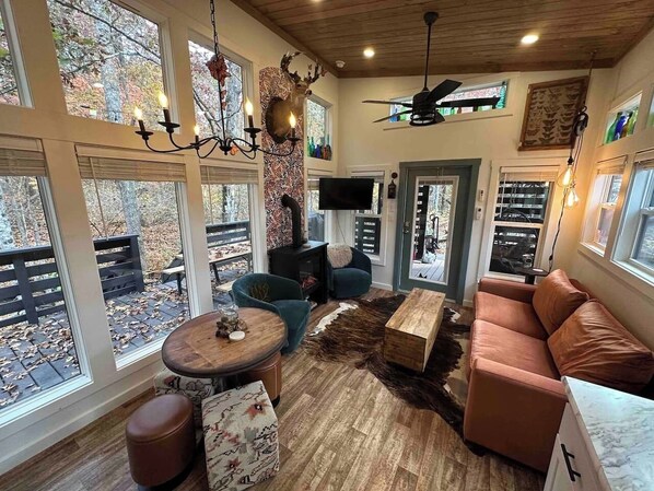 A cozy living room with a fireplace, comfortable seating, and large windows offering a view of the outdoors. - Blueberry Farm Cottage | Hot Tub, Creek, Game Room (Mentone)