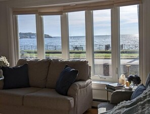 Ein Zimmer mit großem Fenster und Blick auf die Küste, ein beiges Sofa und ein kleiner Tisch mit Büchern und einer Vase.