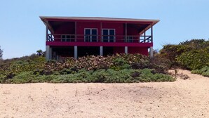 Een rood, twee verdiepingen hoog gebouw met een balkon, omringd door weelderige begroeiing en een zandig voorgrondgebied.