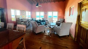 Een ruime woonkamer met een houten plafond, grote ramen met uitzicht op de oceaan, en een eettafel met stoelen.