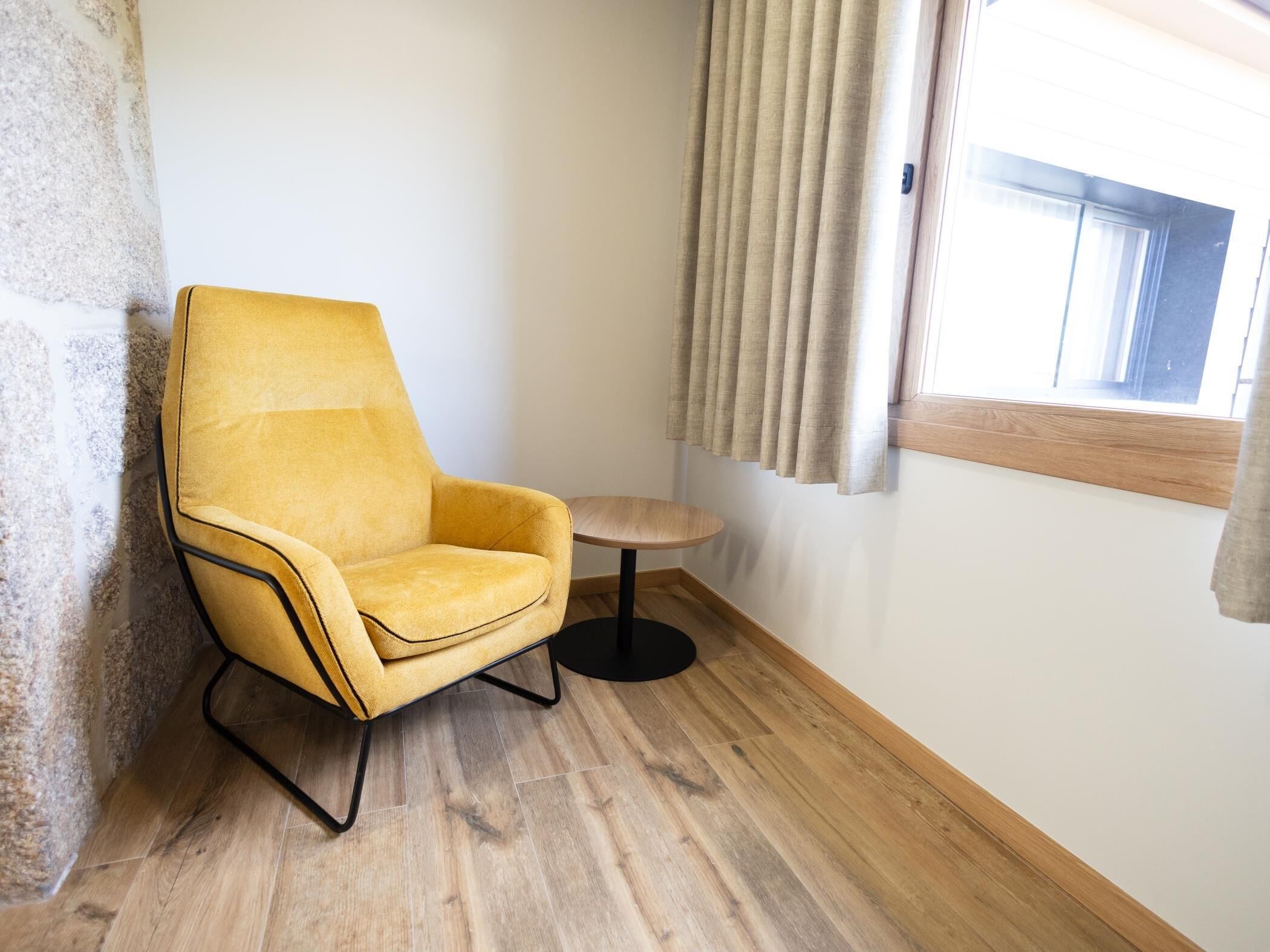 Een hoekzaak met een gele fauteuil, een klein rond tafeltje, een raam met gordijnen en een stenen muur.