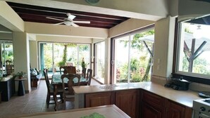 Cozinha com armários de madeira, sala de jantar com mesa e cadeiras, e grandes janelas com vista para vegetação.