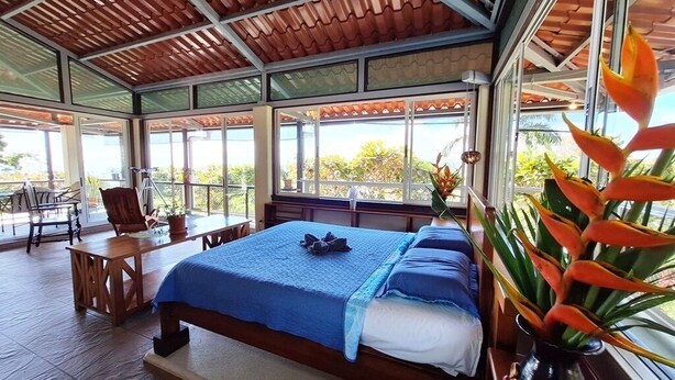 Quarto com cama, escrivaninha e cadeira. Possui janelas amplas e um ventilador de teto.