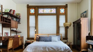 A bedroom with a wooden bed, a desk with books, a piano, and stained glass windows. - Victorian Modern, on the Mississippi River (Minneapolis)