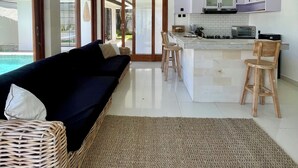 A modern kitchen with white cabinets and a central island, a dining area with wooden chairs, and a black sofa with a woven rug. - 3 br villa across from Thomas beach, Uluwatu . (Pecatu, Bali)
