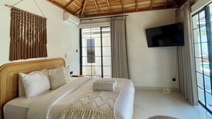 A spacious bedroom with a large bed, wooden ceiling beams, a flat-screen TV, and a view through a large window. - 3 br villa across from Thomas beach, Uluwatu . (Pecatu, Bali)