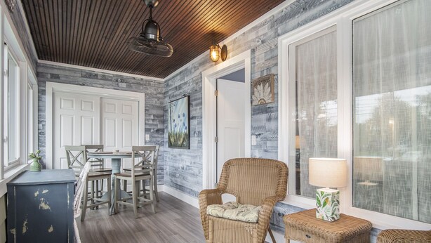 Una habitación acogedora con techo de madera, un comedor con sillas y una mesa, un sillón de mimbre, una mesita auxiliar con una lámpara y un ventanal con cortinas.