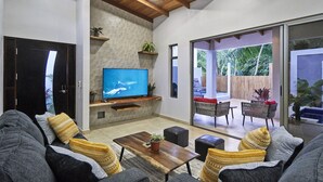 A modern living room with a flat-screen TV, grey sofas, a wooden coffee table, and a sliding glass door leading to an outdoor patio. - 3BD Private Villa Konka with pool  (Cóbano)