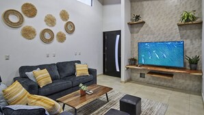 A modern living room with a grey sofa, a wooden coffee table, a flat-screen TV, decorative wall art, and built-in shelves with plants. - 3BD Private Villa Konka with pool  (Cóbano)