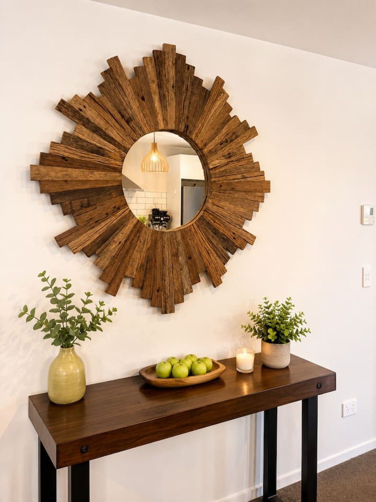 A wall-mounted wooden sunburst mirror with a round mirror below it, flanked by two potted plants and a wooden side table with a bowl of apples and a candle.