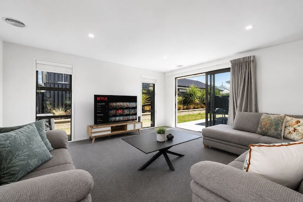 A modern living room with a flat-screen TV, two sofas, a coffee table, and a sliding glass door leading to a garden.