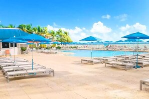 A poolside area with lounge chairs and umbrellas, a swimming pool, and palm trees in the background.