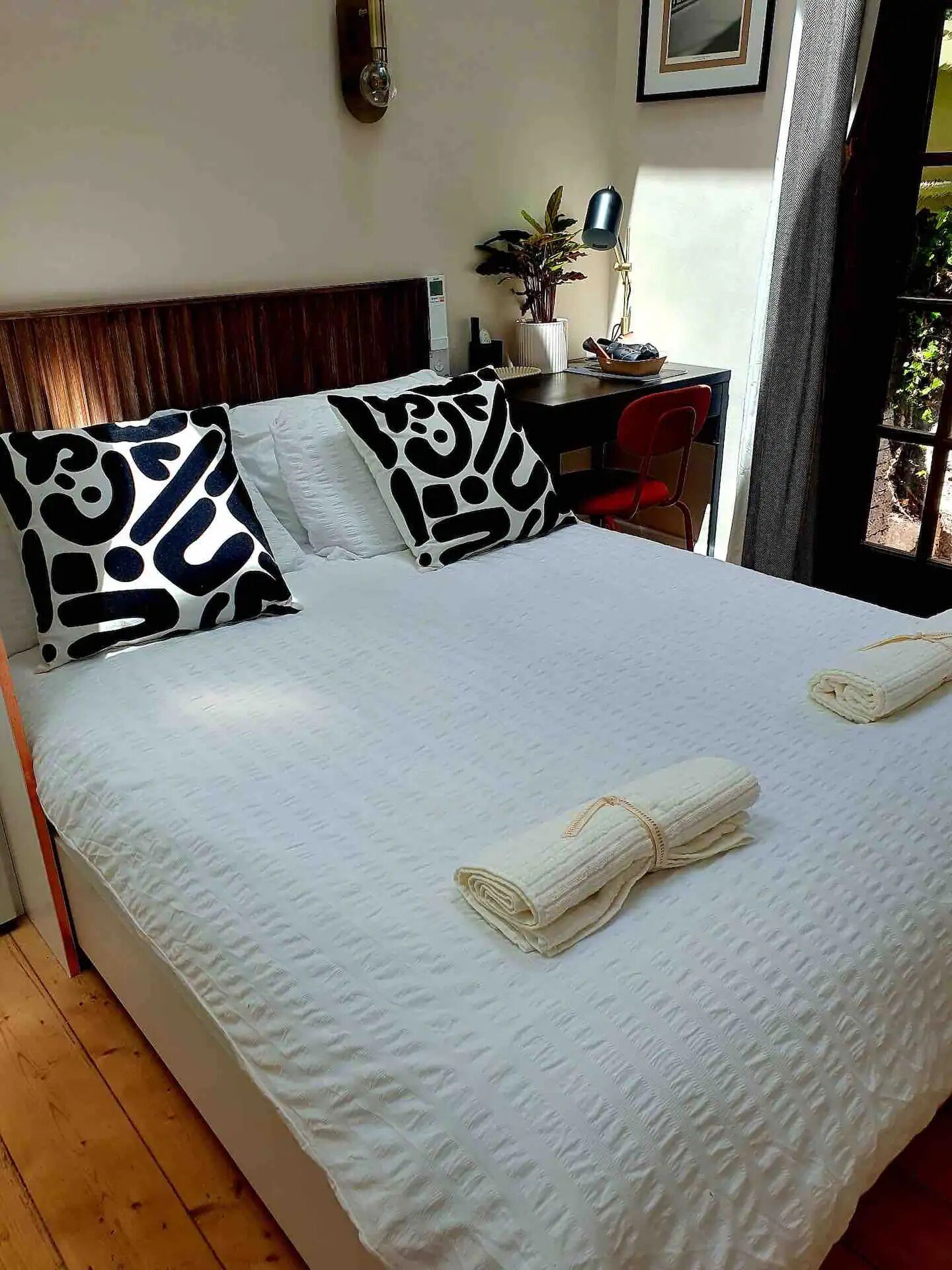 Een netjes opgemaakt bed met wit beddegoed en zwart gestreepte kussens, een bureau met stoel, en een raam met uitzicht op groen.