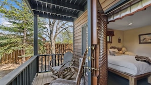 A cozy bedroom with a wooden bed, a small bedside table, and a nightstand. There is a window with a view of trees and a wooden deck outside.