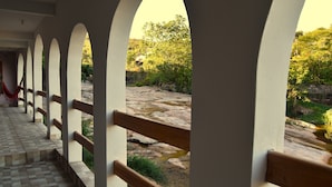 A covered walkway with arches and a wooden railing, leading to a rocky outdoor area with a stream.