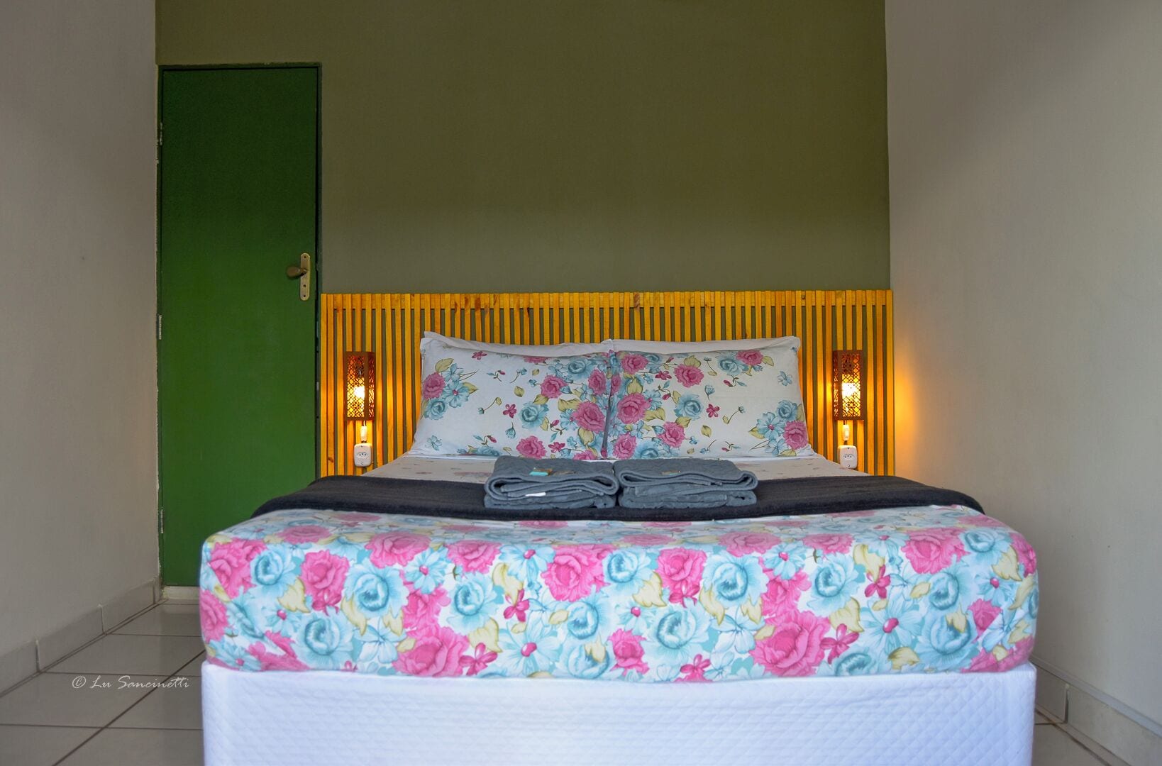 Una cama con ropa de cama estampada, una puerta verde y dos lámparas fijadas a la pared.