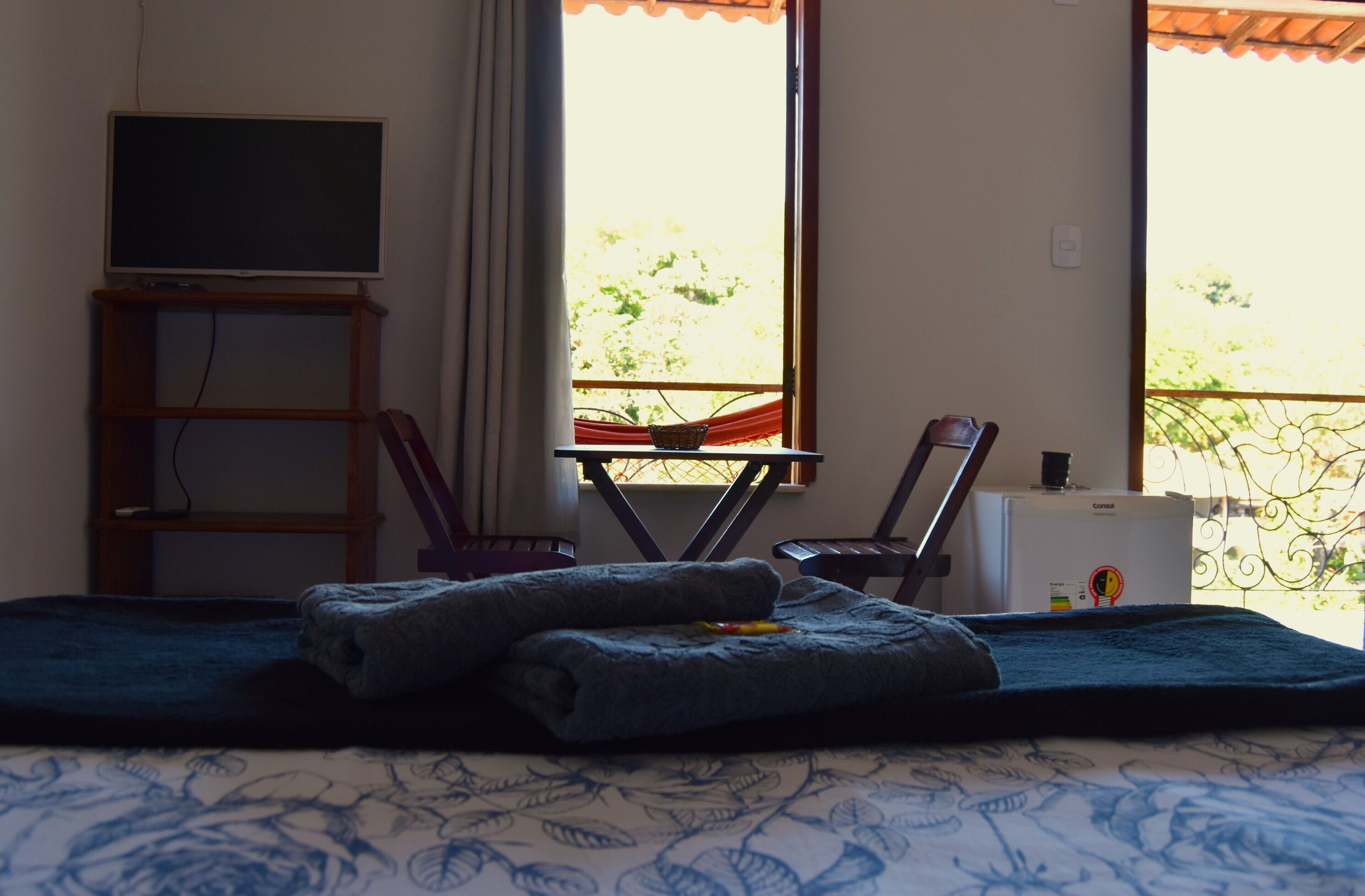 Una habitación con cama, televisor, una mesita con una silla y ventana con vista a follaje.