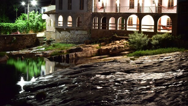 A stone building with arched windows and a small bridge over a reflective water body at night.