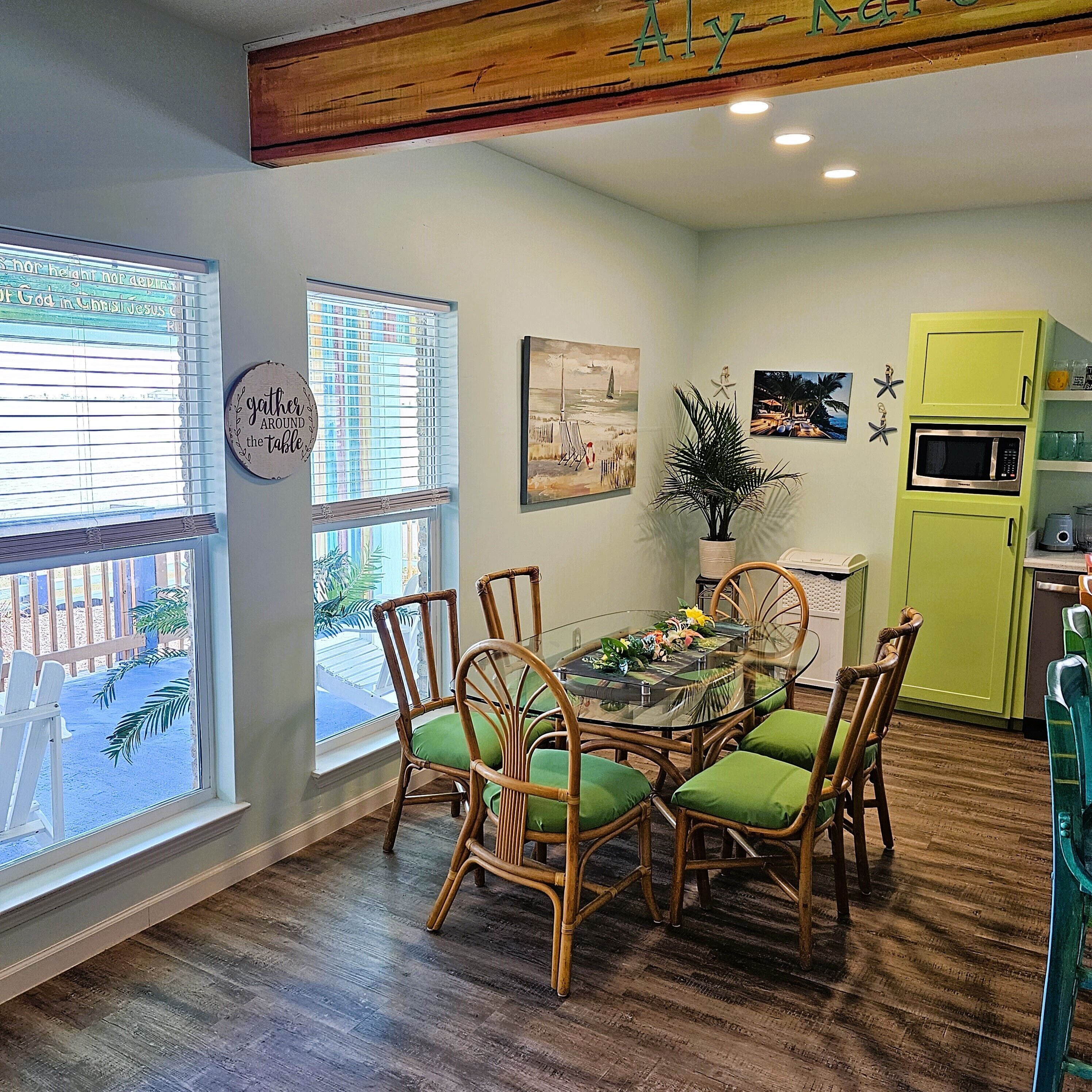 A dining area with a glass table surrounded by wicker chairs with green cushions, wooden flooring, and a view of a deck through large windows.
