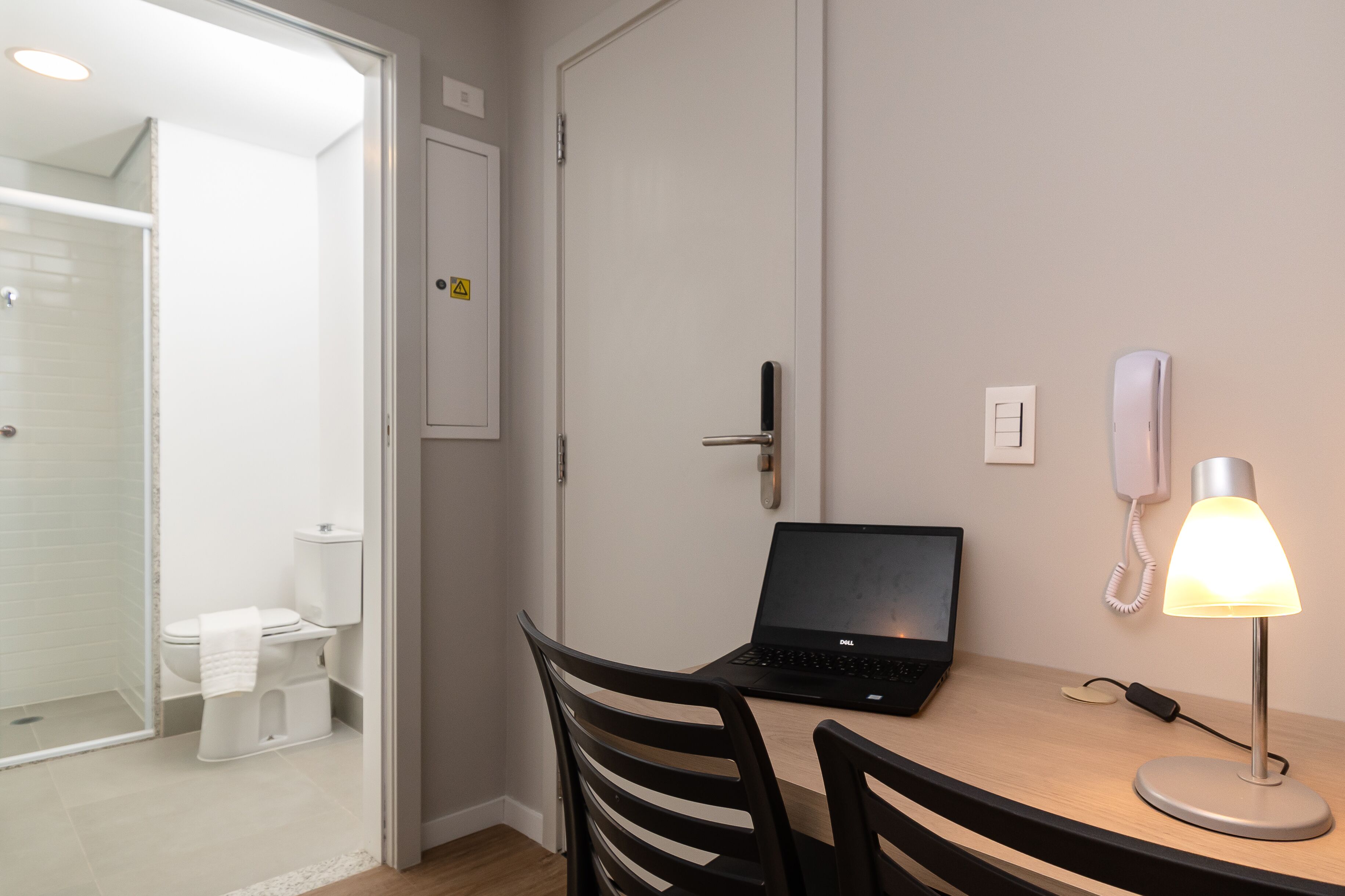 Habitación de hotel con una laptop sobre un escritorio, una lámpara de noche, un teléfono y una puerta que da a un baño.