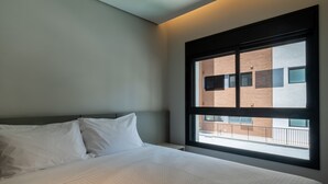 Quarto de hotel com uma grande janela, cama com roupa de cama branca e vista para um edifício.