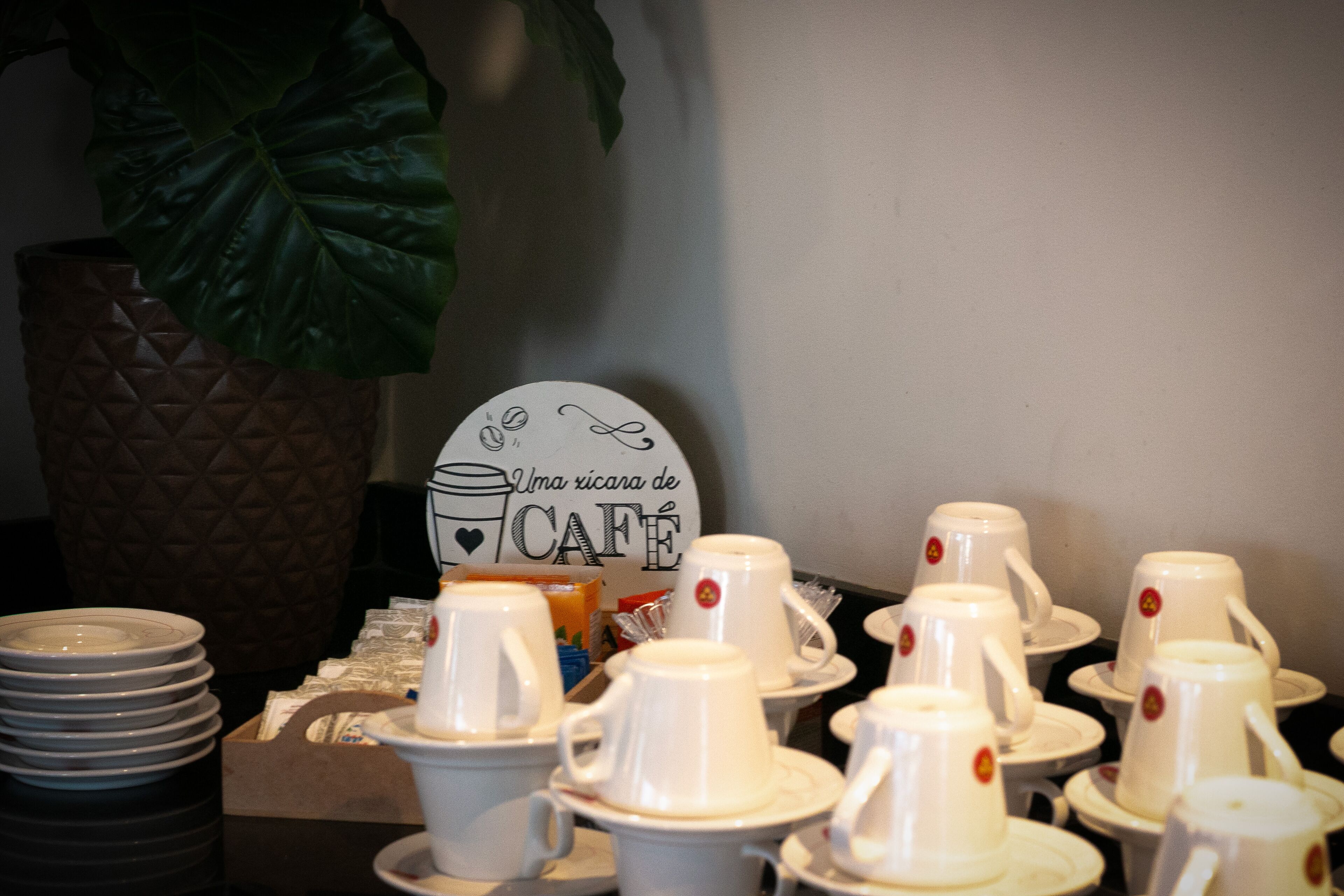 Een tafel met witte kopjes en schotels, een bord met de tekst 'Uma ricaana de CAFE' en een grote plant op de achtergrond.