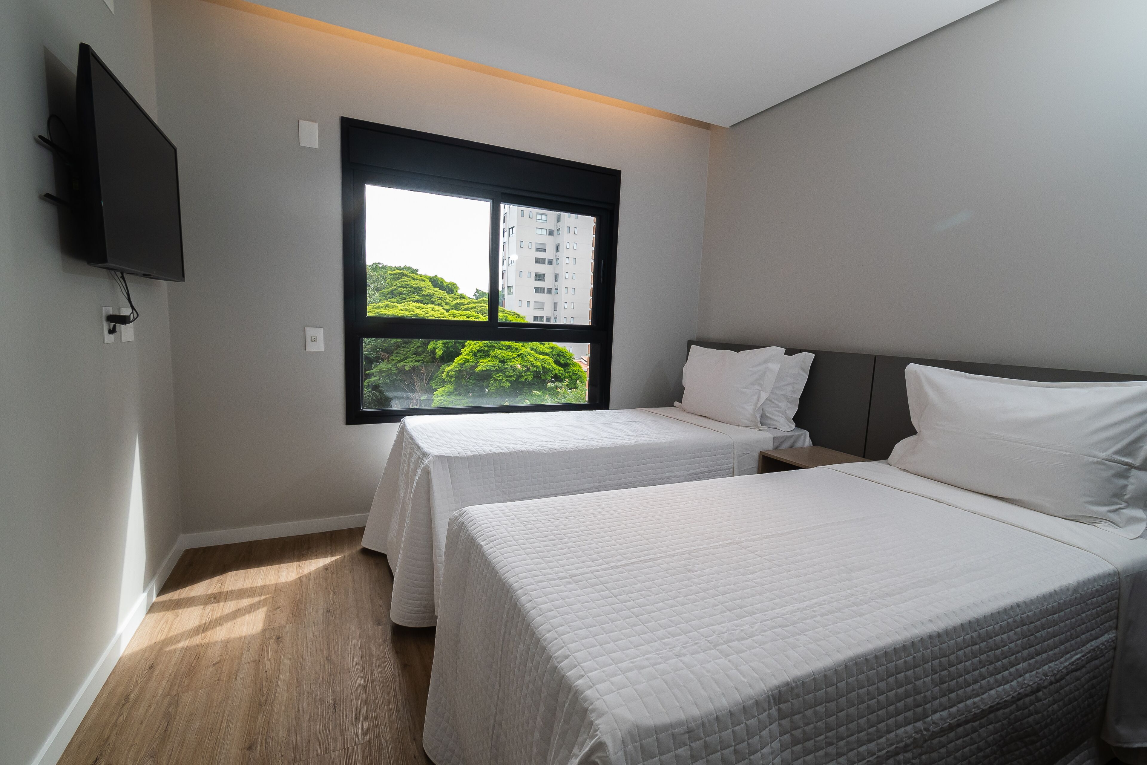 A hotel room with two beds, a TV mounted on the wall, and a window with a view of greenery and a building.