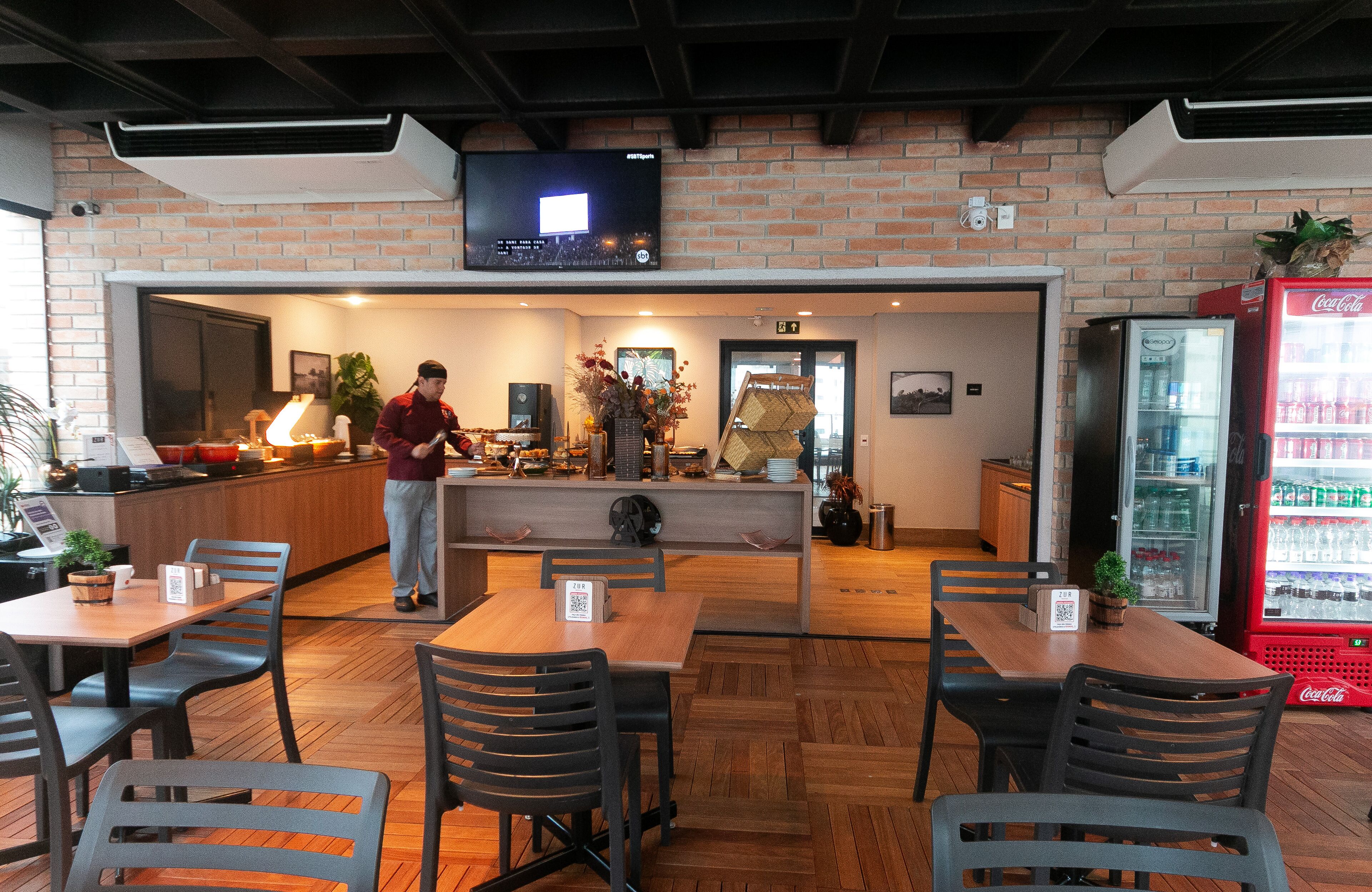 A restaurant with wooden tables and chairs, a TV mounted on the wall, and a Coca-Cola refrigerator.