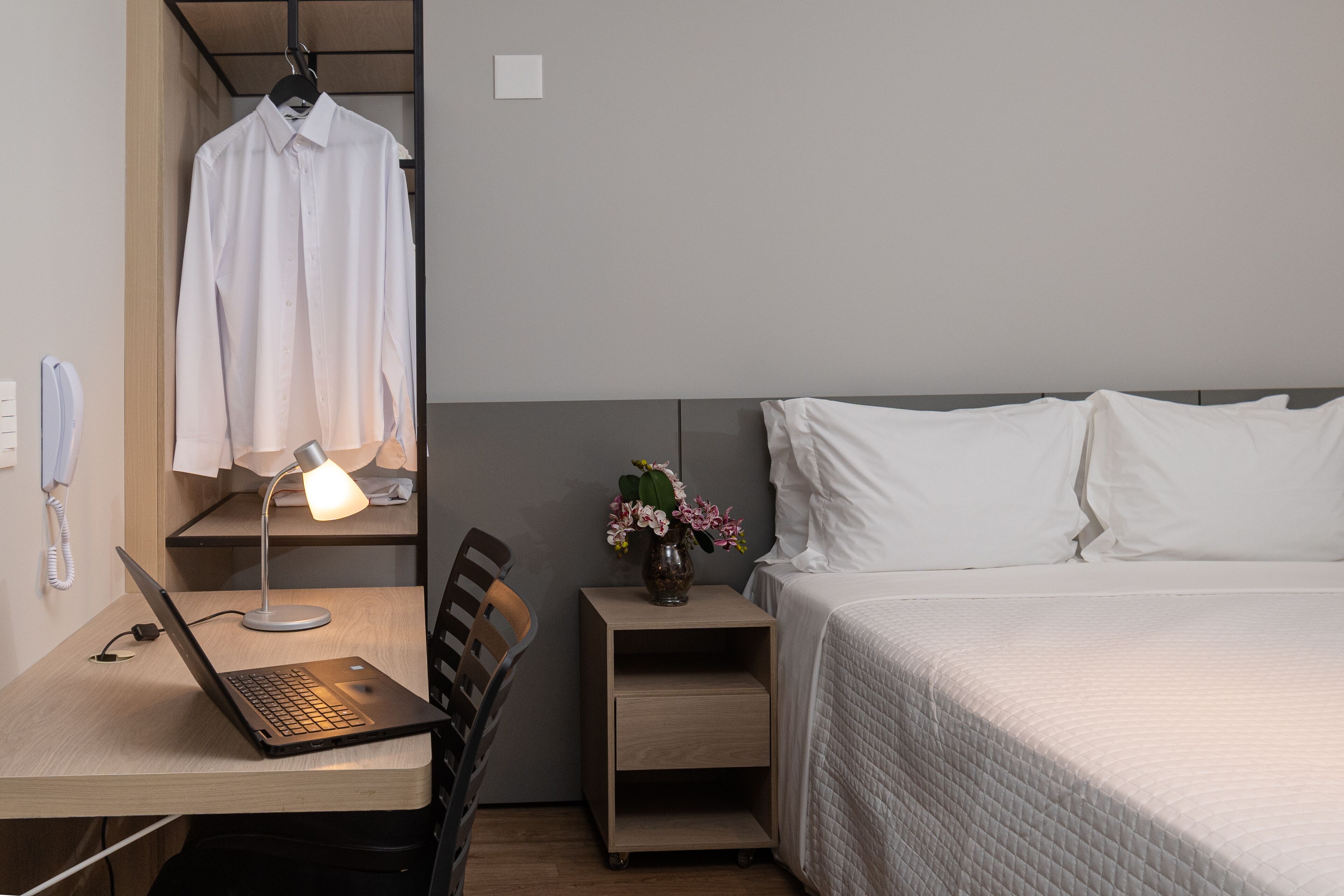 Habitación de hotel con cama, escritorio con una computadora portátil, una silla, mesita de noche con lámpara y flores, y un perchero.